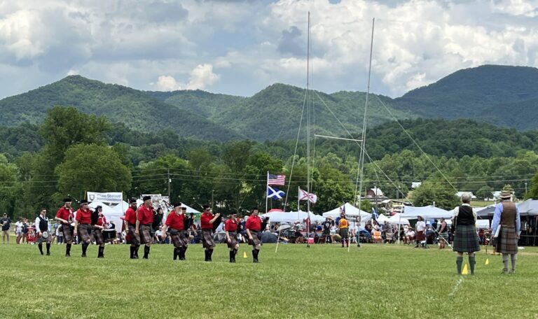 A pipe and drum band marches onto a field with the Great Smoky Mountains in the background.