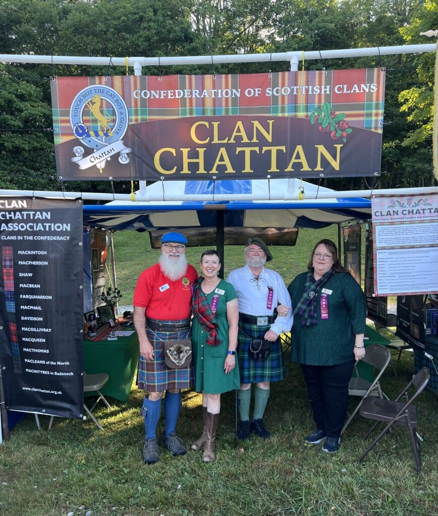Several individuals stand in front of a tent sign that says Clan Chattan at an outdoor festival.